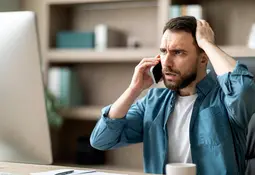 Ein Mann mit besorgtem Gesichtsausdruck hält sich mit einer Hand an den Kopf und telefoniert mit der anderen Hand, im Hintergrund ein Computerbildschirm.