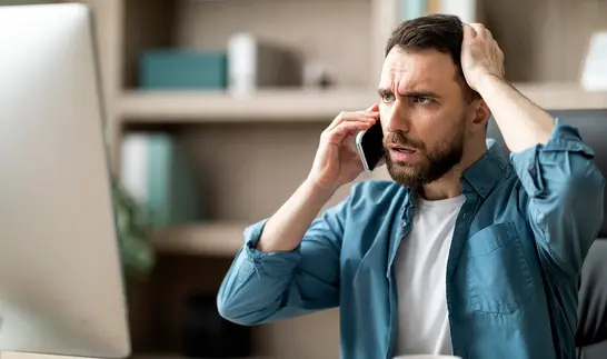 Ein Mann mit besorgtem Gesichtsausdruck hält sich mit einer Hand an den Kopf und telefoniert mit der anderen Hand, im Hintergrund ein Computerbildschirm.