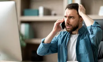 Ein Mann mit besorgtem Gesichtsausdruck hält sich mit einer Hand an den Kopf und telefoniert mit der anderen Hand, im Hintergrund ein Computerbildschirm.