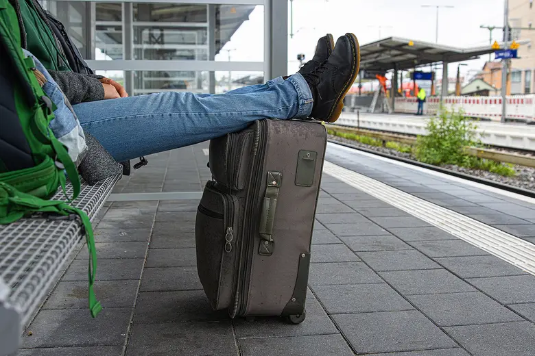 Person sitzt auf einer Bank am Bahnsteig, Füße auf einem Koffer.