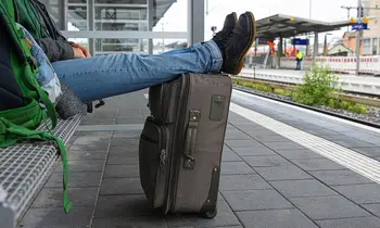 Person sitzt auf einer Bank am Bahnsteig, Füße auf einem Koffer.
