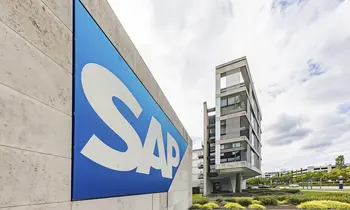 Blaues SAP-Logo mit weißem Schriftzug an einer hellen Betonwand, dahinter ein mehrgeschossiges Gebäude unter bewölktem Himmel.