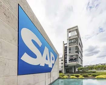 Blaues SAP-Logo mit weißem Schriftzug an einer hellen Betonwand, dahinter ein mehrgeschossiges Gebäude unter bewölktem Himmel.