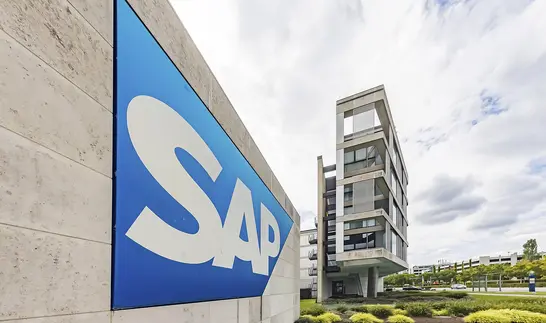 Blaues SAP-Logo mit weißem Schriftzug an einer hellen Betonwand, dahinter ein mehrgeschossiges Gebäude unter bewölktem Himmel.