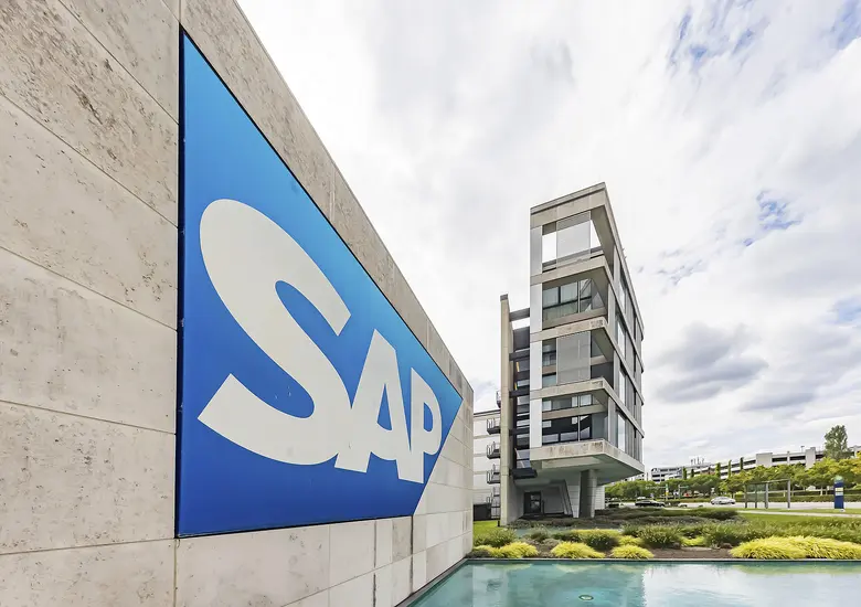 Blaues SAP-Logo mit weißem Schriftzug an einer hellen Betonwand, dahinter ein mehrgeschossiges Gebäude unter bewölktem Himmel.