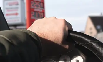 Hand am Lenkrad eines Autos, im Hintergrund unscharf die Preistafel einer Tankstelle.