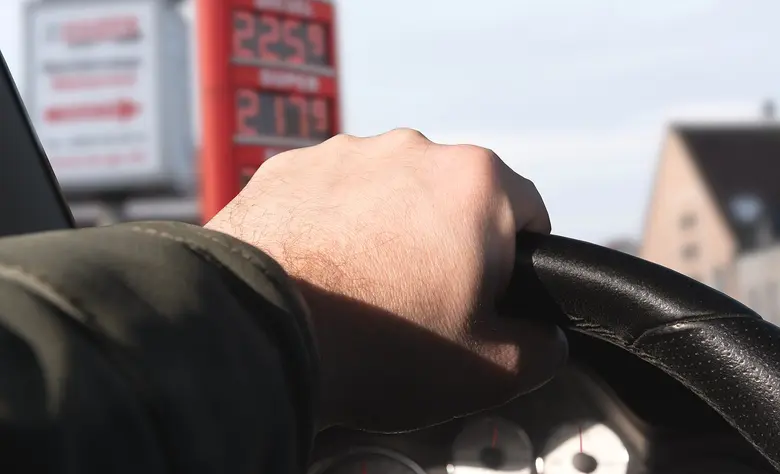Hand am Lenkrad eines Autos, im Hintergrund unscharf die Preistafel einer Tankstelle.