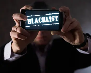 Ein Mann im Anzug hält ein Smartphone mit beiden Händen vor sich, auf dem Display steht in weißen Buchstaben das Wort „Blacklist".