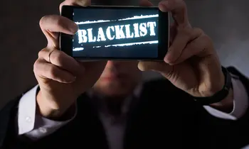 Ein Mann im Anzug hält ein Smartphone mit beiden Händen vor sich, auf dem Display steht in weißen Buchstaben das Wort „Blacklist".