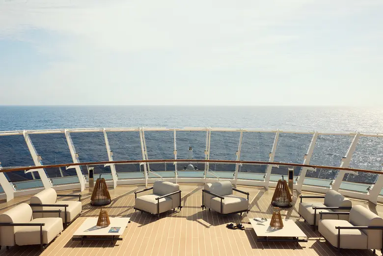 Zu sehen ist die Aussicht von einem Kreuzfahrtschiff auf das offene Meer. An Deck sind Lounge-Möbel platziert.
