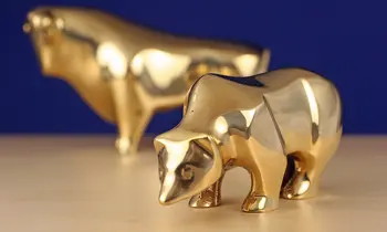 Eine goldene Bärenfigur im Vordergrund und eine goldene Bullenfigur unscharf im Hintergrund auf einer hellen Holzfläche vor blauem Hintergrund.