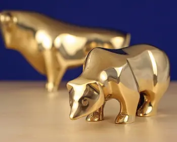 Eine goldene Bärenfigur im Vordergrund und eine goldene Bullenfigur unscharf im Hintergrund auf einer hellen Holzfläche vor blauem Hintergrund.