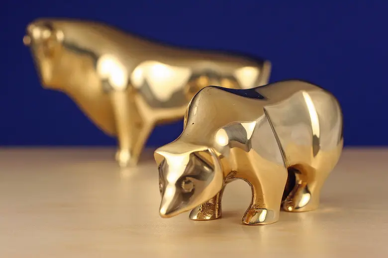 Eine goldene Bärenfigur im Vordergrund und eine goldene Bullenfigur unscharf im Hintergrund auf einer hellen Holzfläche vor blauem Hintergrund.