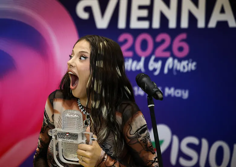 Eine junge Frau mit langen dunklen Haaren hält einen kristallartigen Pokal und reißt den Mund auf, im Hintergrund ein blaues Banner mit der Aufschrift Vienna 2026.