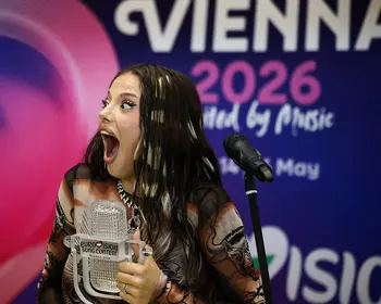 Eine junge Frau mit langen dunklen Haaren hält einen kristallartigen Pokal und reißt den Mund auf, im Hintergrund ein blaues Banner mit der Aufschrift Vienna 2026.