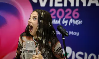 Eine junge Frau mit langen dunklen Haaren hält einen kristallartigen Pokal und reißt den Mund auf, im Hintergrund ein blaues Banner mit der Aufschrift Vienna 2026.