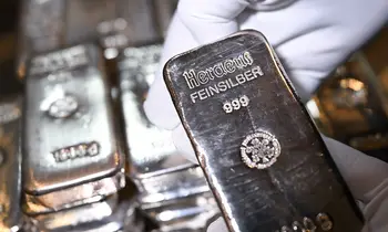 Eine behandschuhte Hand hält einen glänzenden Silberbarren mit der Prägung Heraeus Feinsilber 999, im Hintergrund liegen weitere Barren.