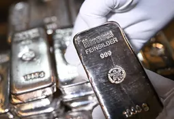 Eine behandschuhte Hand hält einen glänzenden Silberbarren mit der Prägung Heraeus Feinsilber 999, im Hintergrund liegen weitere Barren.