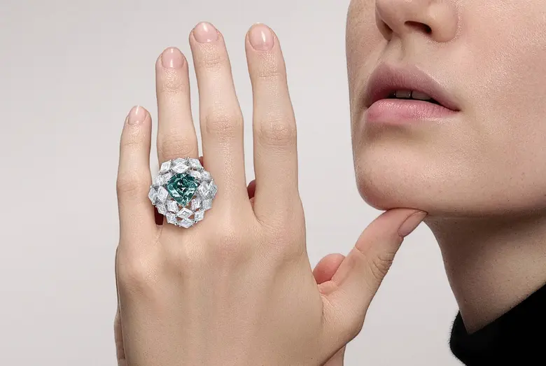 Ein Frau trägt an der linken Hand einen Ring mit einem blaugrünen Diamanten als Mittelstein und vielen weiteren weißen Diamanten.