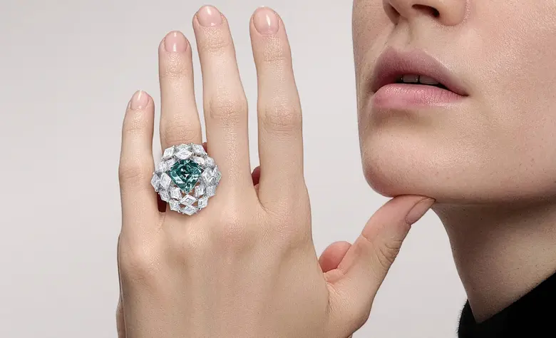 Ein Frau trägt an der linken Hand einen Ring mit einem blaugrünen Diamanten als Mittelstein und vielen weiteren weißen Diamanten.