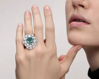 Ein Frau trägt an der linken Hand einen Ring mit einem blaugrünen Diamanten als Mittelstein und vielen weiteren weißen Diamanten.