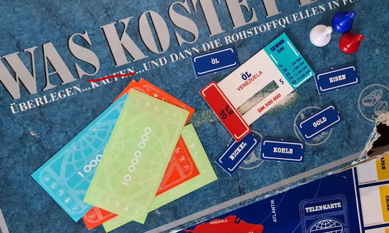 Spielmaterial des Brettspiels „Was kostet die Welt?“ mit Karten für Öl, Kohle, Eisen und Spielgeld auf einer beschädigten Spieleschachtel
