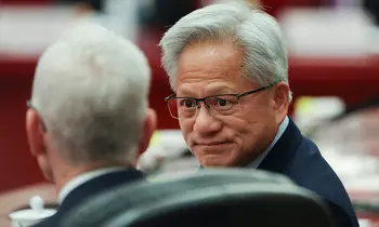 Jensen Huang mit Brille im Gespräch mit einer Person, deren Rücken zur Kamera zeigt, vor unscharfem rotem Hintergrund.
