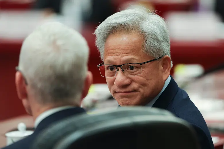 Jensen Huang mit Brille im Gespräch mit einer Person, deren Rücken zur Kamera zeigt, vor unscharfem rotem Hintergrund.