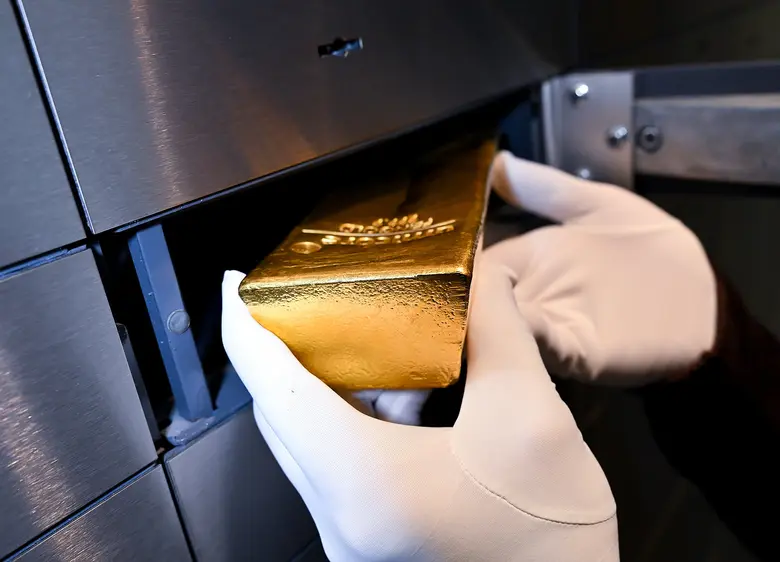 Eine behandschuhte Hand legt einen glänzenden Goldbarren in ein offenes Schließfach aus Metall.