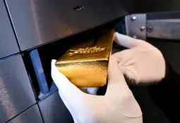 Eine behandschuhte Hand legt einen glänzenden Goldbarren in ein offenes Schließfach aus Metall.