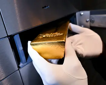 Eine behandschuhte Hand legt einen glänzenden Goldbarren in ein offenes Schließfach aus Metall.