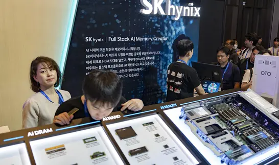 Eine Frau und ein Mann betrachten aufmerksam interaktive Bildschirme und Hardware-Exponate an einem beleuchteten Messestand mit der Aufschrift SK hynix, im Hintergrund weitere Besucher.