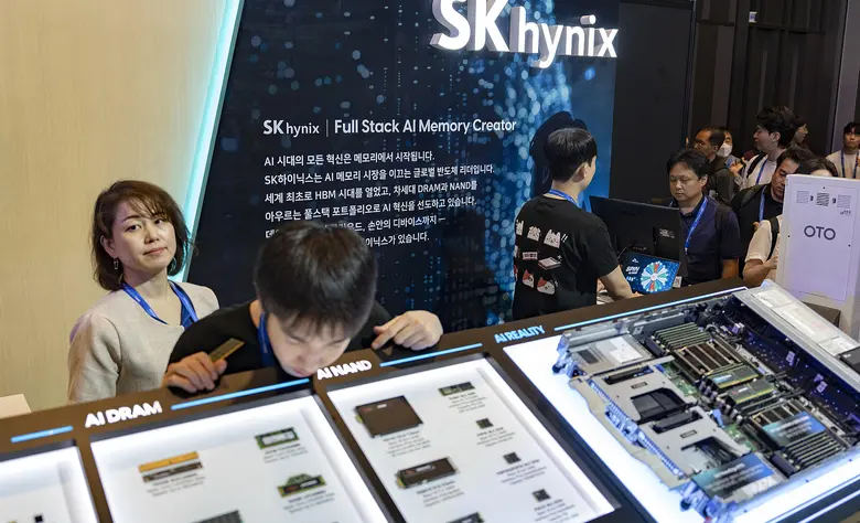 Eine Frau und ein Mann betrachten aufmerksam interaktive Bildschirme und Hardware-Exponate an einem beleuchteten Messestand mit der Aufschrift SK hynix, im Hintergrund weitere Besucher.