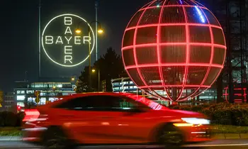 Weiß leuchtendes Bayer-Kreuz und eine große, rot leuchtende Gitterkugel bei Nacht, im Vordergrund ein vorbeifahrendes rotes Auto mit Bewegungsunschärfe.
