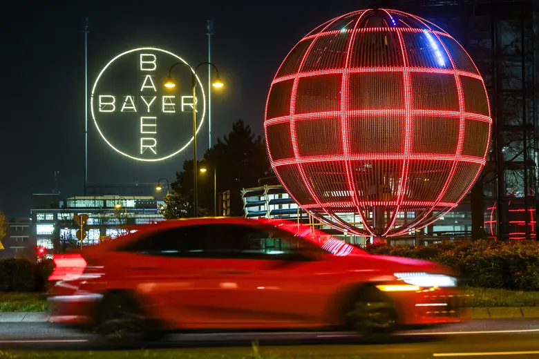 Weiß leuchtendes Bayer-Kreuz und eine große, rot leuchtende Gitterkugel bei Nacht, im Vordergrund ein vorbeifahrendes rotes Auto mit Bewegungsunschärfe.