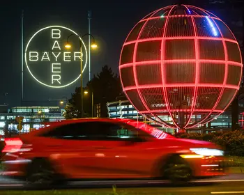 Weiß leuchtendes Bayer-Kreuz und eine große, rot leuchtende Gitterkugel bei Nacht, im Vordergrund ein vorbeifahrendes rotes Auto mit Bewegungsunschärfe.