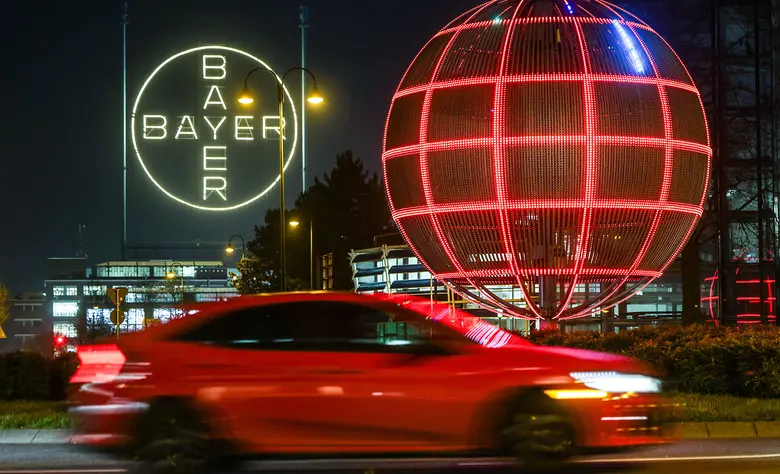Weiß leuchtendes Bayer-Kreuz und eine große, rot leuchtende Gitterkugel bei Nacht, im Vordergrund ein vorbeifahrendes rotes Auto mit Bewegungsunschärfe.