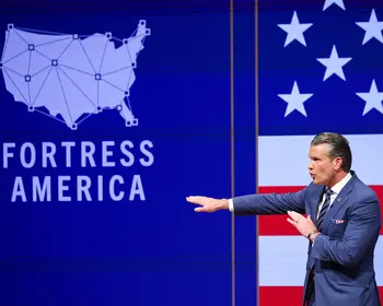 Ein Mann in blauem Anzug streckt beim Sprechen einen Arm nach links aus, hinter ihm sind eine blaue Leinwand mit einer Karte der USA und dem Schriftzug Fortress America sowie eine US-Flagge zu sehen.