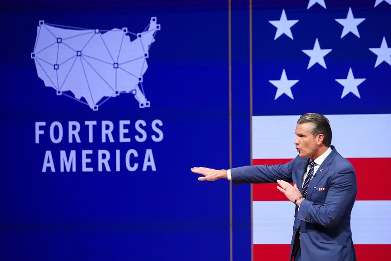Ein Mann in blauem Anzug streckt beim Sprechen einen Arm nach links aus, hinter ihm sind eine blaue Leinwand mit einer Karte der USA und dem Schriftzug Fortress America sowie eine US-Flagge zu sehen.