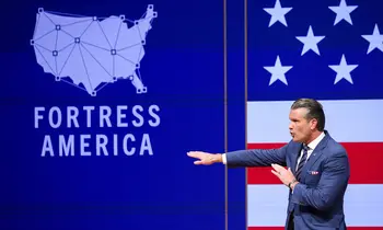 Ein Mann in blauem Anzug streckt beim Sprechen einen Arm nach links aus, hinter ihm sind eine blaue Leinwand mit einer Karte der USA und dem Schriftzug Fortress America sowie eine US-Flagge zu sehen.