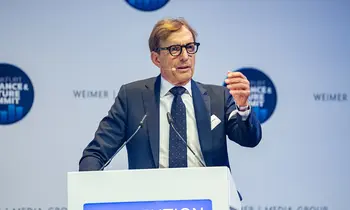 Ein Mann mit Brille, dunkelblauem Anzug und Krawatte steht an einem Rednerpult mit Mikrofonen. Während seiner Rede gestikuliert er mit erhobener rechter Hand.