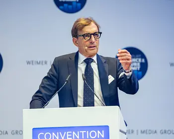 Ein Mann mit Brille, dunkelblauem Anzug und Krawatte steht an einem Rednerpult mit Mikrofonen. Während seiner Rede gestikuliert er mit erhobener rechter Hand.
