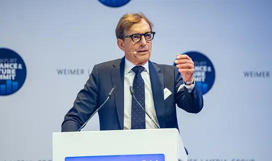 Ein Mann mit Brille, dunkelblauem Anzug und Krawatte steht an einem Rednerpult mit Mikrofonen. Während seiner Rede gestikuliert er mit erhobener rechter Hand.