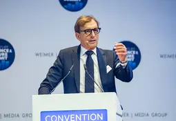 Ein Mann mit Brille, dunkelblauem Anzug und Krawatte steht an einem Rednerpult mit Mikrofonen. Während seiner Rede gestikuliert er mit erhobener rechter Hand.