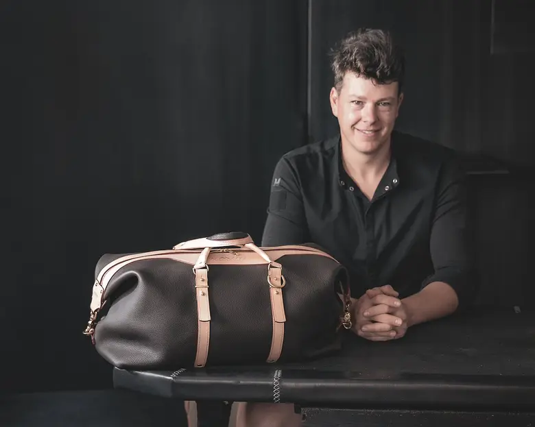Ein lächelnder Lederhandwerker sitzt hinter einer dunkelbraunen Reisetasche mit hellbraunen Lederriemen und goldfarbenen Metallbeschlägen.