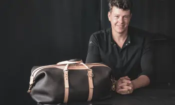 Ein lächelnder Lederhandwerker sitzt hinter einer dunkelbraunen Reisetasche mit hellbraunen Lederriemen und goldfarbenen Metallbeschlägen.