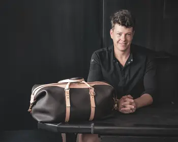 Ein lächelnder Lederhandwerker sitzt hinter einer dunkelbraunen Reisetasche mit hellbraunen Lederriemen und goldfarbenen Metallbeschlägen.