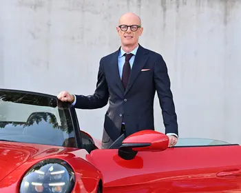 Ein glatzköpfiger Mann mit dunkler Brille, dunkelblauem Anzug und hellblauem Hemd steht lächelnd neben einem roten Sportwagen mit geöffneter Tür.