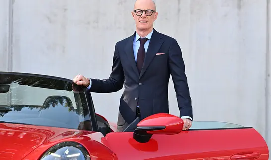 Ein glatzköpfiger Mann mit dunkler Brille, dunkelblauem Anzug und hellblauem Hemd steht lächelnd neben einem roten Sportwagen mit geöffneter Tür.