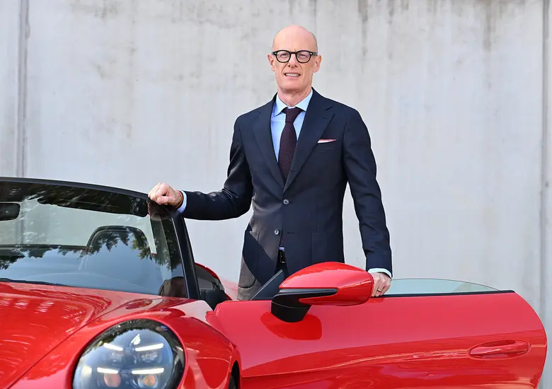 Ein glatzköpfiger Mann mit dunkler Brille, dunkelblauem Anzug und hellblauem Hemd steht lächelnd neben einem roten Sportwagen mit geöffneter Tür.